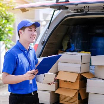 delivery driver or courier in blue uniform and hat loading cardboard boxes from car trunk with clipboard for logistics company.